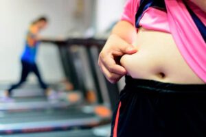 Uma mulher segurando a barriga enquanto a outra na esteira faz exercícios para queimar gordura