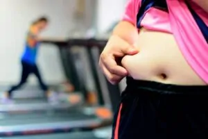 Uma mulher segurando a barriga enquanto a outra na esteira faz exercícios para queimar gordura