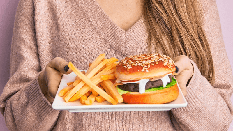 imagem de uma mulher segurando um prato com hambúrguer e batata frita, alimentos que aumentam o colesterol
